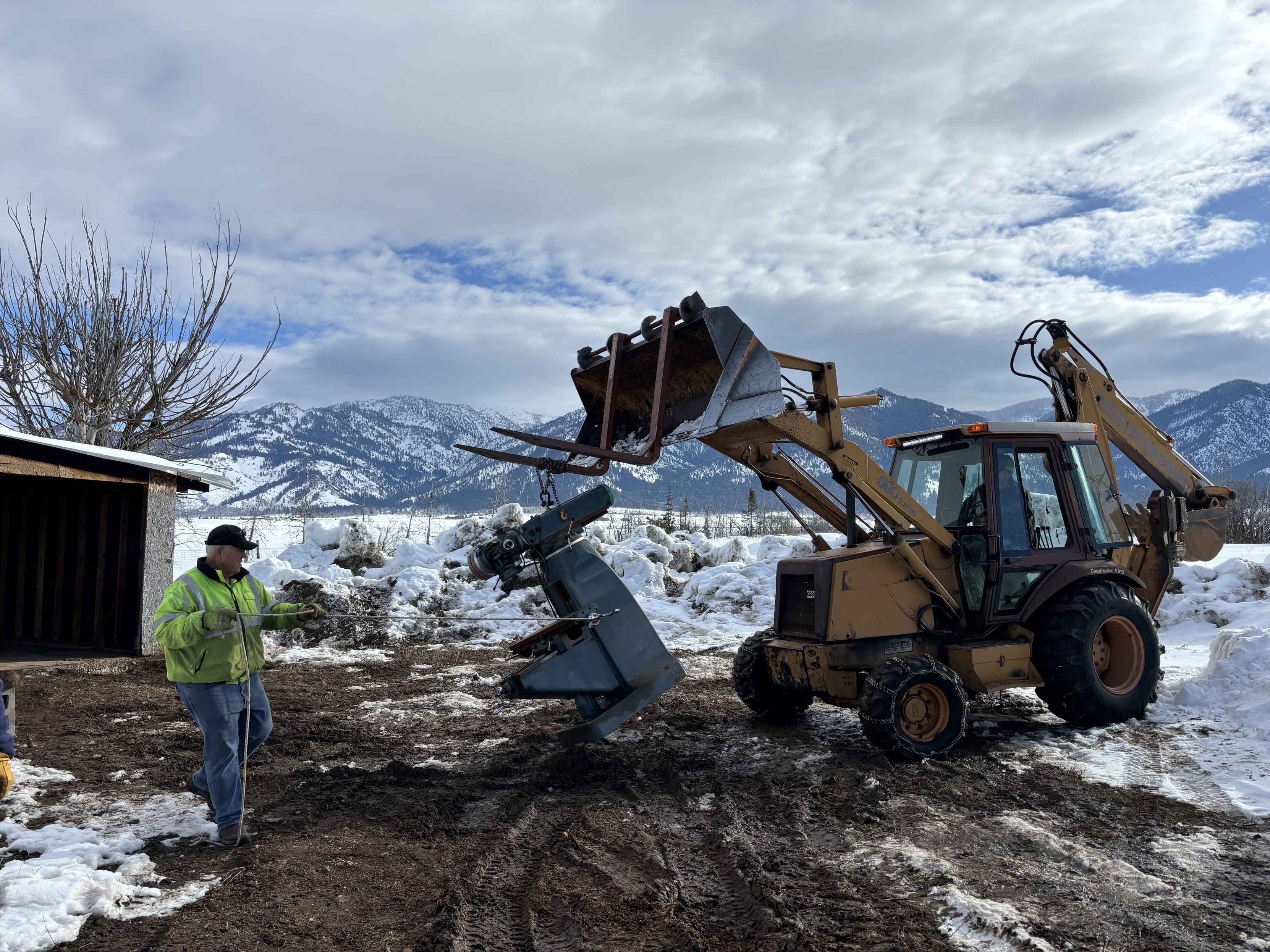 Backhoe lifting a mill while a worker watches. Mountains with snow in the background.